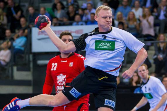 Ein männlicher Handballspieler in einem weißen Trikot springt und bereitet sich auf den Wurf vor, während ein Verteidiger in einem roten Trikot versucht, ihn zu blockieren. Zuschauer beobachten das Geschehen im Hintergrund.