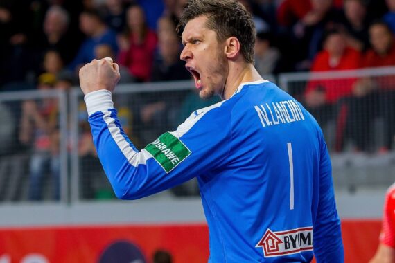 Ein männlicher Handballspieler in einem blauen Trikot mit der Aufschrift Alilovic und der Nummer 1 auf dem Rücken reckt die Faust in die Höhe und jubelt auf einem Hallenplatz, wobei im Hintergrund Zuschauer und andere Spieler zu sehen sind.