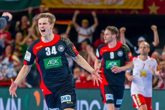 Ein männlicher Handballspieler in einem schwarz-roten Trikot mit der Nummer 34 jubelt mit ausgestreckten Armen auf einem Spielfeld, während Mannschaftskameraden und ein Gegner in der Nähe stehen und im Hintergrund jubelnde Fans zu sehen sind.