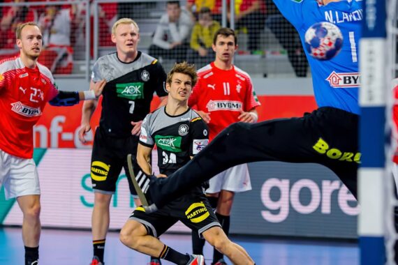 Ein Handballspieler in einem schwarz-grauen Trikot bereitet sich auf einen Schuss vor, während der Torwart in Blau den Ball abwehrt; andere Spieler in Rot und Schwarz beobachten das Geschehen aufmerksam.