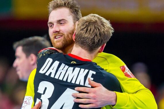 Zwei männliche Sportler in Sportuniformen umarmen sich nach einem Spiel. Der Spieler, der der Kamera zugewandt ist, trägt einen Bart, während der andere die DAHMKE 34 auf der Rückseite seines Trikots trägt. Sie scheinen zu feiern oder sich gegenseitig zu trösten.
