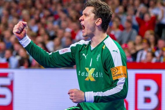 Ein männlicher Handballspieler in einem grünen Trikot jubelt mit geballter Faust und offenem Mund und scheint zu schreien. Er trägt eine orangefarbene Kapitänsbinde, und im Hintergrund ist eine Zuschauermenge zu sehen.