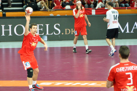 Ein Handballspieler in einem roten Trikot bereitet sich während eines Spiels auf den Wurf vor, während andere Spieler, ebenfalls in Rot und einer in Weiß, auf dem Spielfeld zu sehen sind. Die Zuschauer schauen von den Tribünen im Hintergrund zu.