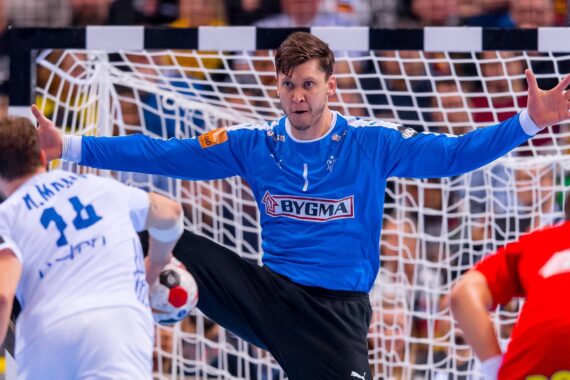 Ein Handballtorwart in einem blauen Trikot mit weit gespreizten Armen und Beinen versucht, einen Schuss abzuwehren, während er einem Spieler in Weiß gegenübersteht, der sich anschickt, den Ball zu werfen. Das Tornetz und andere Spieler sind im Hintergrund zu sehen.