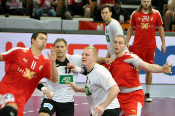 Vier Handballspieler, zwei in Rot und zwei in Weiß, kämpfen auf einem Hallenplatz aktiv um den Ball. Andere Spieler und Zuschauer sind im Hintergrund zu sehen. Die Szene ist dynamisch und intensiv.