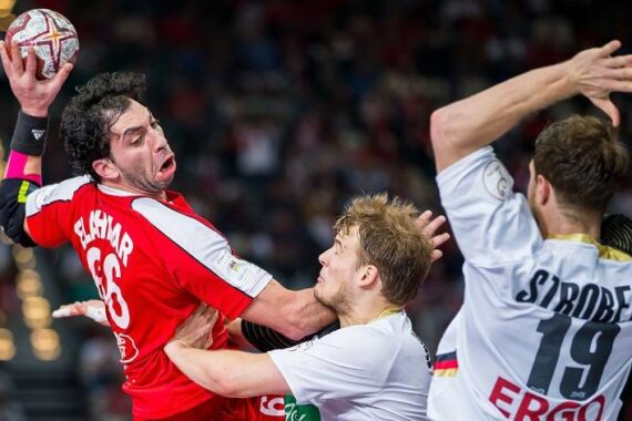 Ein Handballspieler in einem roten Trikot springt zum Schuss, während zwei Gegner in weißen Trikots den Schuss abwehren, wobei einer versucht, den Schuss zu blockieren und der andere nach dem Spieler greift. Die Aktion findet in einer Halle statt.