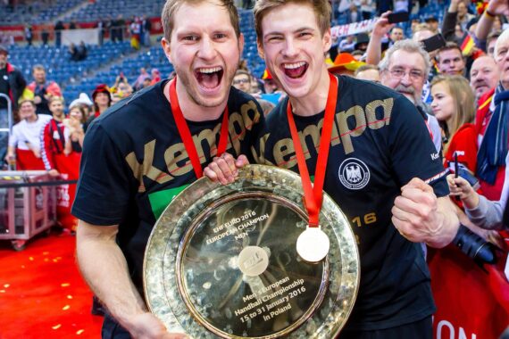 Zwei jubelnde männliche Athleten in schwarzen Kempa-Hemden und mit Goldmedaillen halten eine große Meisterschaftstrophäe und feiern mit jubelnden Fans in einem Stadion.