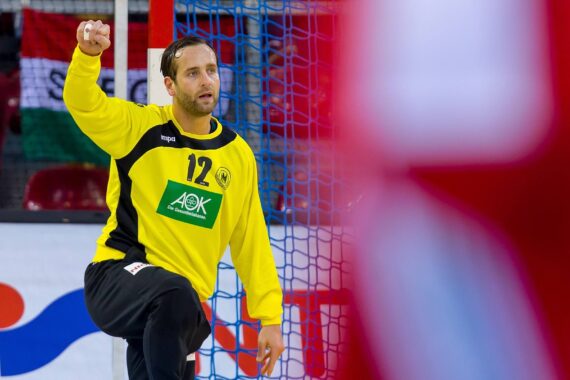 Ein männlicher Handballtorwart in einem gelben Trikot mit der Nummer 12 hebt während eines Spiels in der Nähe des Torpfostens die Faust, während im Vordergrund ein verschwommener Spieler in Rot zu sehen ist.