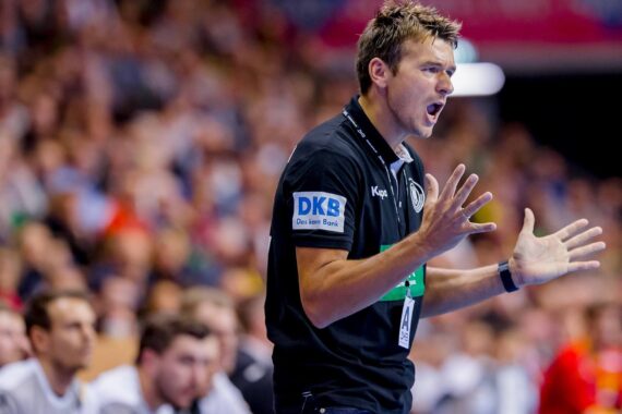 Ein männlicher Handballtrainer steht am Spielfeldrand, gestikuliert mit offenen Händen und ruft, wirkt intensiv und konzentriert. Unscharfe Spieler und Zuschauer füllen den Hintergrund.