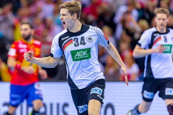 Ein männlicher Handballspieler, der ein weiß-schwarzes Trikot mit der Nummer 34 trägt, jubelt mit geballter Faust auf dem Spielfeld, während im Hintergrund Mannschaftskameraden und Gegner zu sehen sind.