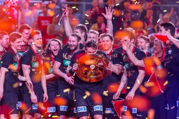 Eine Gruppe jubelnder Athleten feiert auf einer Bühne, wobei einer ein großes Trophäenschild in der Hand hält. Um sie herum fällt orangefarbenes Konfetti, und die Teamkollegen lächeln, klatschen und schwenken Fahnen in der festlichen Atmosphäre.