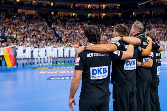 Vier deutsche Handballer stehen mit den Armen um die Schultern vor einer Reihe von Spielern und einer deutschen Fahne auf dem Spielfeld, während im Hintergrund eine Menschenmenge die Arena füllt.