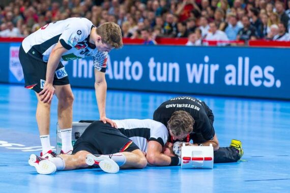 Ein Handballspieler liegt verletzt auf dem Spielfeld, während ein Sanitäter und ein Mannschaftskamerad sich um ihn kümmern. Der Hintergrund zeigt eine Menschenmenge und ein hellblaues Werbebanner mit deutschem Text.