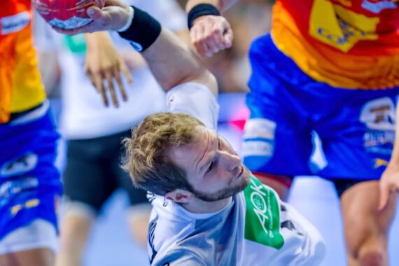 Ein männlicher Handballspieler in einem weißen Trikot springt mit einem roten Ball in der Hand nach vorne, während zwei Gegner in blauen und gelben Trikots versuchen, ihn während eines intensiven Spiels zu blockieren.
