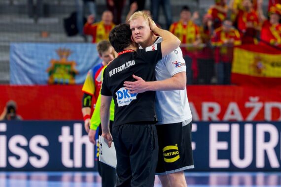 Zwei männliche Handballspieler umarmen sich auf dem Spielfeld, wobei der eine den anderen tröstet. Im Hintergrund sieht man jubelnde Fans in Rot und Gelb und Nationalflaggen, darunter die spanische, was auf eine Sportveranstaltung hindeutet.