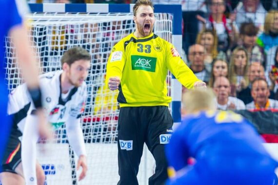 Ein Handballtorwart in einem gelben Trikot mit der Nummer 33 feiert mit ausgestreckten Armen und offenem Mund, während im Hintergrund Spieler und Zuschauer zu sehen sind.