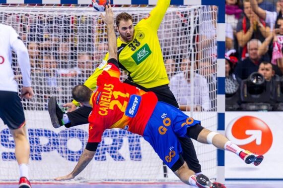 Ein Handballtorwart in Gelb blockiert während eines Spiels einen Schuss eines Spielers in einem rot-blauen Trikot in der Luft, während die Zuschauer von der Tribüne aus zusehen.