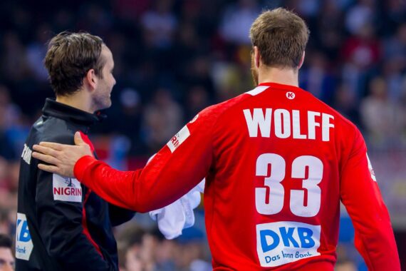 Zwei männliche Sportler in rot-schwarzen Mannschaftsuniformen stehen zusammen in einer Arena. Der eine, mit WOLFF 33 auf dem Rücken, hat den Arm um seinen Mannschaftskameraden gelegt. Der Hintergrund ist mit Zuschauern unscharf.