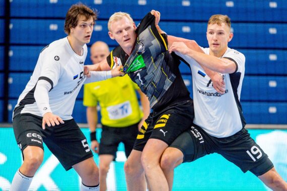 Drei männliche Handballspieler kämpfen um den Ball. Zwei Spieler in weißen Trikots verteidigen sich gegen einen Spieler in schwarz-gelber Uniform, der sich an seinem Trikot festhält. Der Hintergrund zeigt blaue Sitze und einen Schiedsrichter in Gelb.