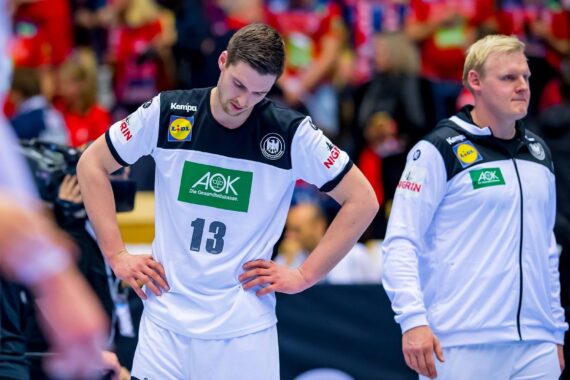 Zwei männliche Handballspieler in weißen und schwarzen Trikots stehen enttäuscht auf dem Spielfeld. Ein Spieler steht mit gesenktem Kopf und Händen in den Hüften, während der andere neben ihm hergeht. Die Zuschauer in Rot sind im Hintergrund verschwommen.