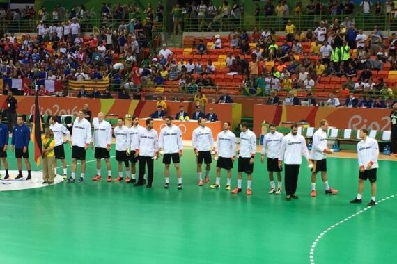 Eine Herren-Handballmannschaft in weißen Trikots steht in einer Reihe auf einem grünen Spielfeld und blickt auf das Publikum in einer Halle. Die Zuschauer füllen die orangefarbenen und gelben Sitze im Hintergrund.