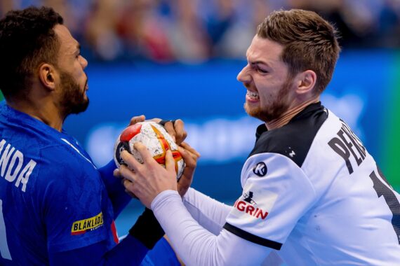 Zwei männliche Handballspieler aus gegnerischen Mannschaften kämpfen intensiv um die Kontrolle über den Ball, den sie während eines Spiels fest in der Hand halten. Ein Spieler trägt Blau, der andere Weiß und Schwarz. Die Szene ist actionreich und angespannt.