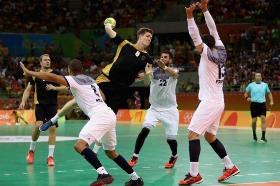 Ein Handballspieler in einem schwarz-gelben Trikot springt, um den Ball zu werfen, während drei Verteidiger in weißen Trikots versuchen, ihn während eines Spiels zu blockieren; im Hintergrund sind ein Schiedsrichter und Zuschauer zu sehen.