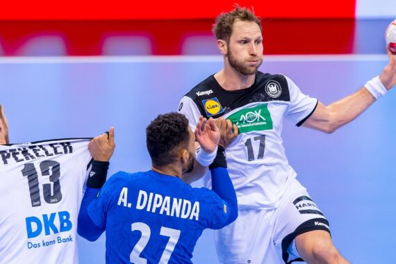 Ein Handballspieler in einem weißen Trikot springt, um den Ball zu werfen, während ein Verteidiger in einem blauen Trikot das Hemd eines anderen weiß gekleideten Spielers festhält. Die Szene spielt sich während eines Spiels auf einem Hallenplatz ab.