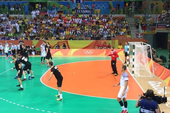 Ein Handballspiel der Männer bei den Olympischen Spielen, bei dem eine Mannschaft angreift und die andere in Tornähe verteidigt. Die Zuschauer füllen die Tribünen, und auf dem Spielfeld sind die olympischen Ringe zu sehen. Ein Kameramann filmt das Geschehen von der Seitenlinie aus.