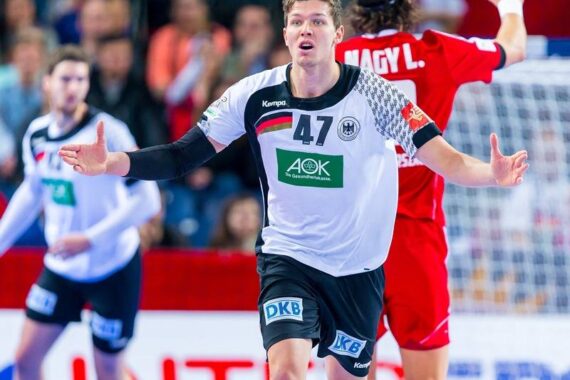 Ein männlicher Handballspieler in einem weiß-schwarzen Trikot mit der Nummer 47 breitet seine Arme auf dem Spielfeld aus. Ein Spieler in einem roten Trikot und andere verschwommene Spieler erscheinen im Hintergrund. Auf der Tribüne sind Zuschauer zu sehen.
