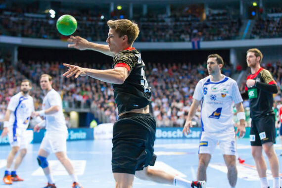 Ein Handballspieler in einem schwarzen Trikot springt, um den Ball zu werfen, während seine Gegner in weißen Trikots auf einem hell erleuchteten Hallenplatz mit einer großen Menschenmenge im Hintergrund zusehen.