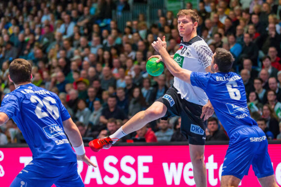 Ein Handballspieler in einem weißen Trikot springt, um den Ball zu werfen, während zwei Gegner in blauen Trikots versuchen, ihn zu blockieren. Das Spiel findet in einer mit Zuschauern gefüllten Halle statt.