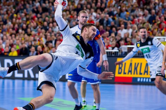Ein Handballspieler in Weiß befindet sich in der Luft und ist im Begriff, den Ball zu werfen, während Spieler in Blau und Zuschauer den Hintergrund einer Sporthalle füllen.