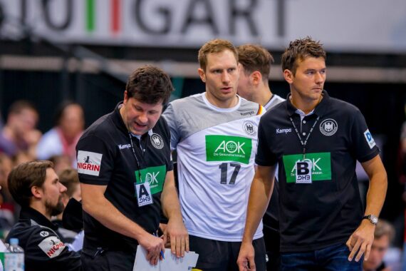 Drei Männer in schwarz-weißer Sportkleidung stehen in einer Sporthalle zusammen. Einer von ihnen, der die Nummer 17 trägt, scheint ein Athlet zu sein, während die anderen Trainer oder Mitarbeiter zu sein scheinen. Die Menge schaut aus dem Hintergrund zu.