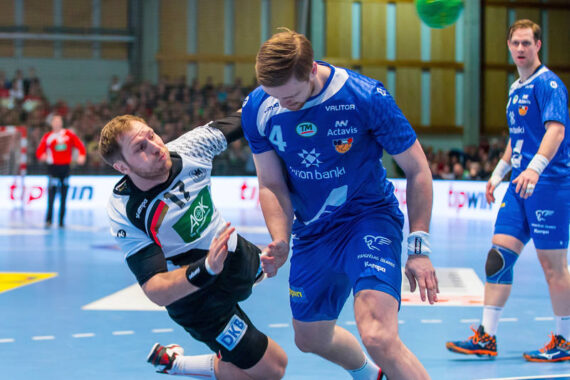 Ein Handballspieler in einem schwarz-weißen Trikot fällt seitwärts, als er mit einem Spieler in einem blauen Trikot zusammenstößt. Der grüne Ball ist in der Luft, und im Hintergrund sind andere Spieler und Zuschauer zu sehen.