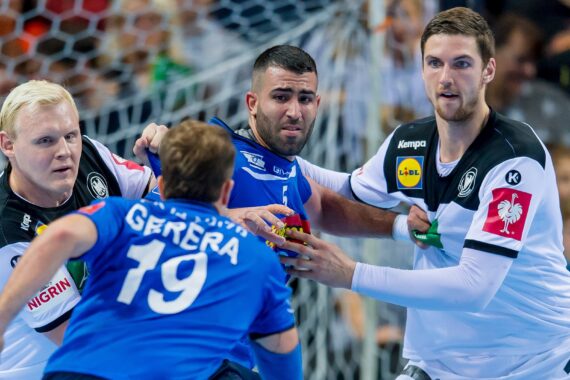 Drei männliche Handballspieler in Aktion während eines Spiels, wobei zwei Spieler in weißen Trikots gegen einen Spieler in einem blauen Trikot verteidigen, der den Ball hält. Im Hintergrund ist das Publikum zu sehen.