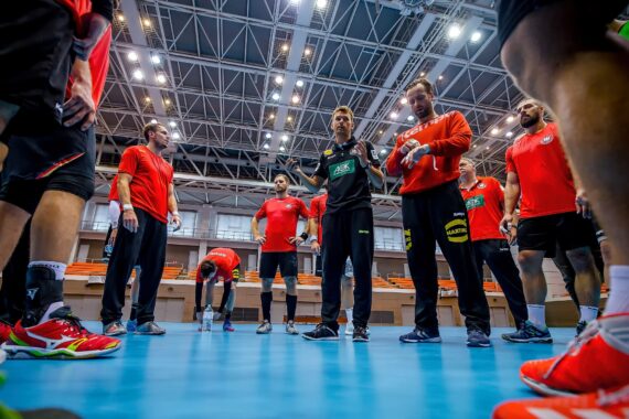 Eine Gruppe männlicher Athleten in Sportkleidung steht in einer Halle zusammen und hört einem Trainer während einer Mannschaftsbesprechung oder einer Trainingseinheit unter hellem Hallenlicht zu.