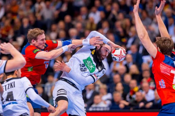 Ein Handballspieler in einem weißen Trikot versucht zu schießen, wird aber von einem Verteidiger in einem roten Trikot geblockt. Ein anderer Verteidiger hebt die Arme, und die Zuschauer im Hintergrund schauen zu. Die Szene ist intensiv und actionreich.