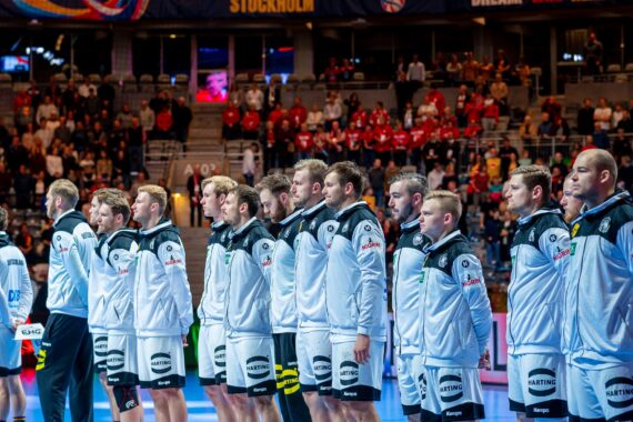 Eine Herren-Handballmannschaft steht in einer Reihe auf dem Spielfeld, trägt weiße Jacken und blickt während einer Zeremonie vor dem Spiel nach vorne, während im Hintergrund ein großes Publikum zu sehen ist.