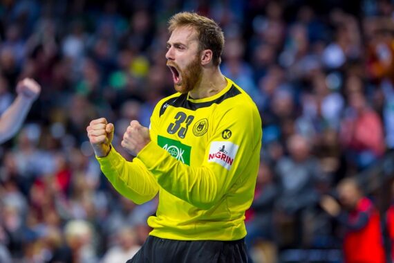 Ein männlicher Handballspieler in einem gelben Trikot mit der Nummer 33 ballt die Fäuste und jubelt, während im Hintergrund eine unscharfe Menschenmenge zu sehen ist.