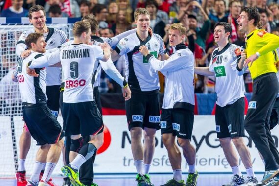Eine Gruppe männlicher Handballspieler in weißen und schwarzen Trikots feiert gemeinsam auf einem Hallenplatz, lächelt und umarmt sich, während die Fans auf der Tribüne im Hintergrund zusehen.