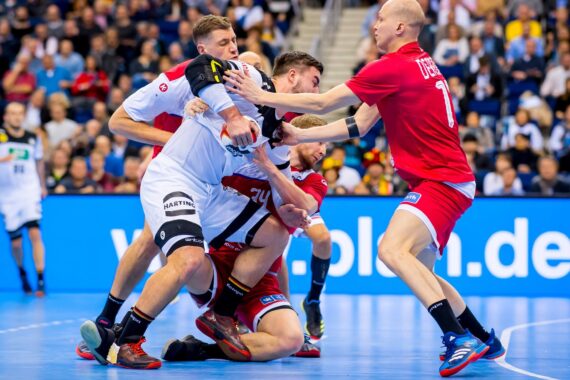 Vier männliche Handballspieler, zwei in weißen und zwei in roten Trikots, sind während eines Spiels auf einem blauen Hallenplatz in einen körperlichen Kampf um den Ball verwickelt. Die Zuschauer füllen die Tribünen im Hintergrund.