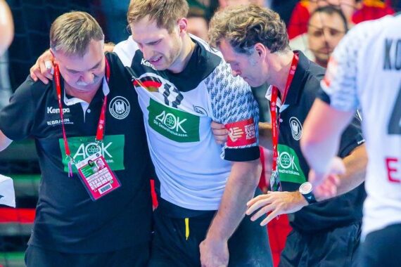 Ein männlicher Sportler im Trikot der deutschen Mannschaft wird von zwei Mitarbeitern, die ihn unter den Armen stützen, vom Spielfeld getragen. Der Spieler scheint verletzt zu sein und zuckt vor Schmerzen.