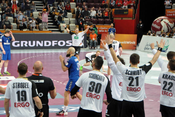 Ein Handballspiel ist in einer Halle im Gange. Die Spieler in weißen Trikots feiern im Vordergrund, während die Spieler in blauen Trikots auf dem Spielfeld reagieren. Die Zuschauer schauen von den Tribünen im Hintergrund zu.