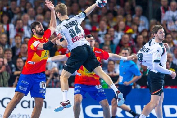Ein Handballspieler in einem weißen Trikot springt während eines Spiels zum Schuss, während er von Spielern in roten Trikots verteidigt wird, während im Hintergrund eine Zuschauermenge zusieht.