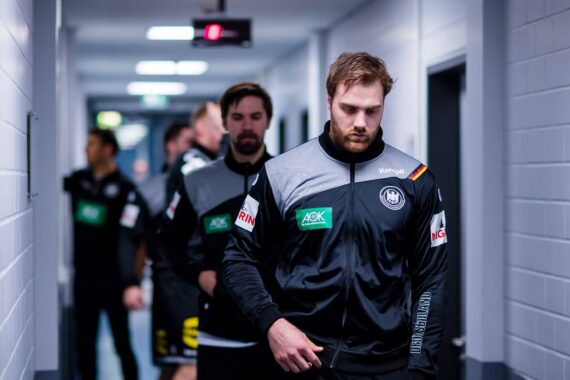 Eine Gruppe männlicher Athleten in passenden schwarzen und grauen Trainingsanzügen geht einen Gang entlang, konzentriert und ernst, auf dem Weg zu einer Veranstaltung oder einem Wettkampf.