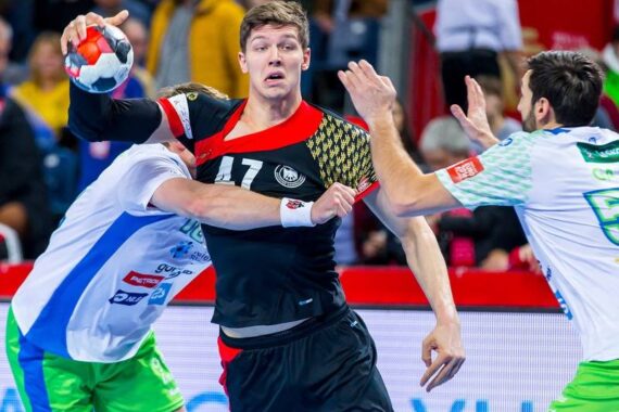 Ein Handballspieler in einem schwarz-roten Trikot versucht, den Ball zu werfen, während er während eines Spiels von zwei Spielern in weiß-grünen Trikots eng verteidigt wird.