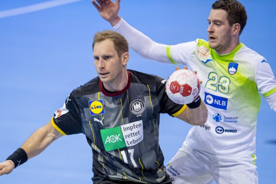 Zwei männliche Handballspieler in Aktion; der eine in einem schwarzen Trikot hält den Ball, der andere in einem weiß-grünen Trikot verteidigt ihn auf einem blauen Hallenplatz.