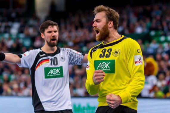 Zwei männliche Handballspieler feiern leidenschaftlich auf dem Spielfeld; der eine im gelben Trikot mit der Nummer 33 ballt die Faust und schreit, während der andere im weißen Trikot mit der Nummer 10 im Hintergrund die Arme hochreißt.