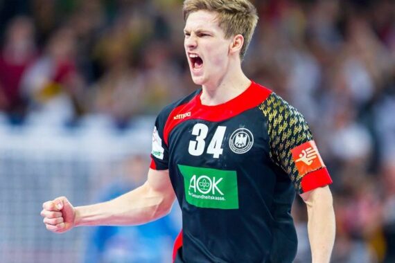 Ein männlicher Handballspieler, der ein schwarz-rotes Trikot mit der Nummer 34 trägt, ballt während eines Spiels die Faust und schreit leidenschaftlich, während im Hintergrund eine verschwommene Menschenmenge zu sehen ist.